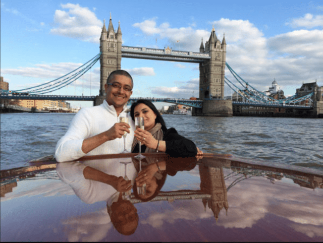 Couple enjoy champagne aboard the luxury Thames Limo boat cruise in front of Tower Bridge, London.