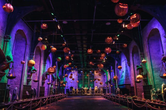 The Great Hall at Warner Bros Studio Tour London – The Making of Harry Potter is pictured at Halloween. Pumpkins appear to float near the ceiling and dramatic lighting in purple and green showcase the hall's tall arched windows.