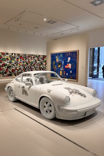 Daniel Arsham's unique white Porsche art installation at Moco Museum London, surrounded by colourful contemporary paintings. The striking sculpture contrasts with vibrant wall art, showcasing Moco Museum's eclectic modern art collection in a bright and spacious gallery setting.