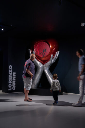 Visitors at Moco Museum London admiring Lorenzo Quinn's art installation, featuring large silver hands holding a red heart with intricate designs. The contemporary art piece attracts a diverse audience, including a family with a child, as they explore the immersive exhibition space.