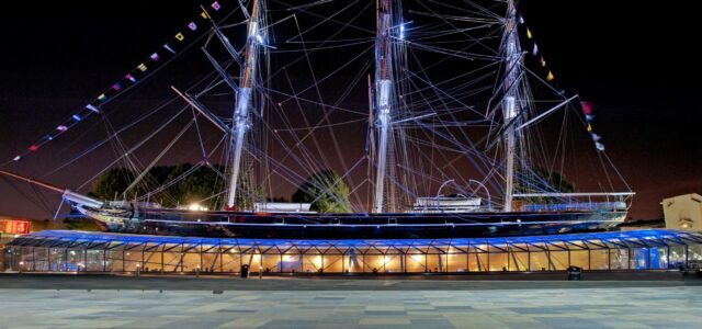 The full length of the Cutty Sark tea clipper ship is pictured from the side, illuminated at night.