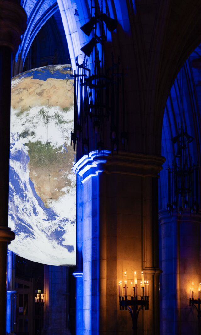 Gaia by Luke Jerram is pictured suspended inside Southwark Cathedral, which is dark apart from some carefully positioned blue uplights that illuminate the Cathedral's dramatic architecture. Gaia is a large recreation of Earth, created to scale using NASA data and imagery. The globe can be seen peeking between two columns, with candles lighting up the foreground of the dark interior of the Cathedral at night.